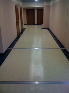 Korytarz z beżową wykładziną podłogową, przecięty dwoma metalowymi listwami dylatacyjnymi, widok na drzwi po obu stronach korytarza.